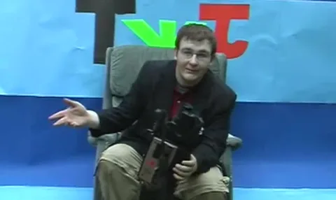 Jon in a blazer on the TWJ set, holding a camcorder