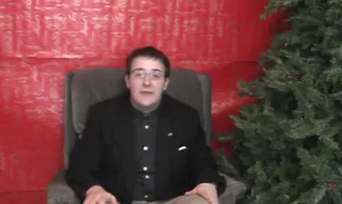 Jon in a dark blazer sitting in an armchair by a red wall and a tree